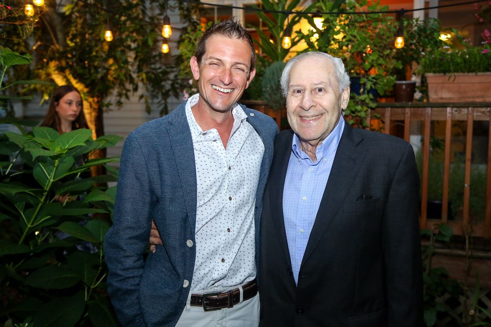 Two men pose together at an evening event with greenery and warm lighting in the background.
