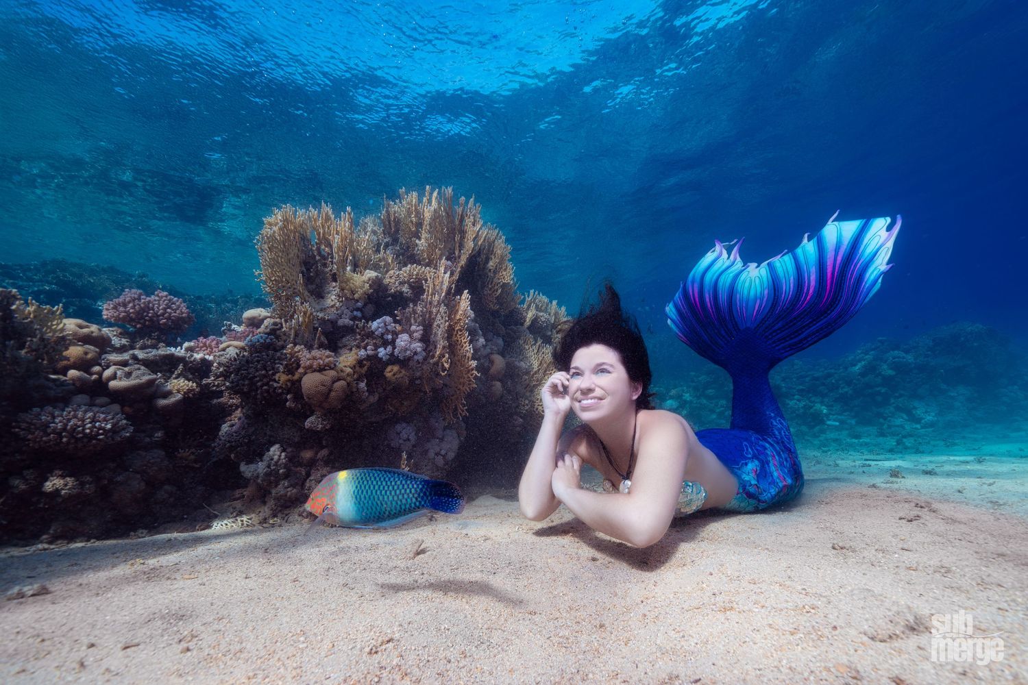 A magical underwater scene of a mermaid reclining on the ocean floor near coral in crystal clear blue waters.