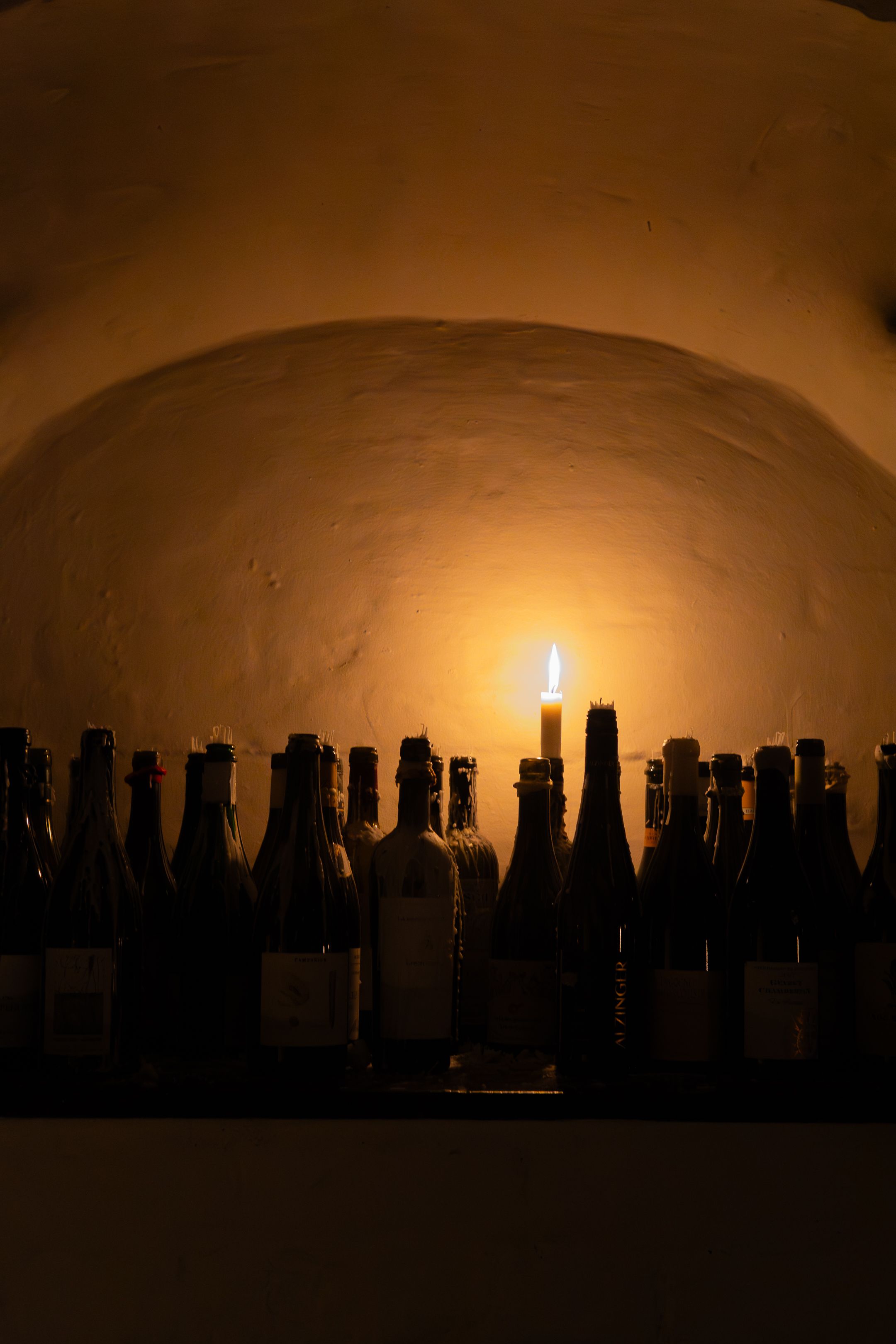 Silhouetted wine bottles against a warm backlit glow with a single candle creating an atmospheric cellar scene.