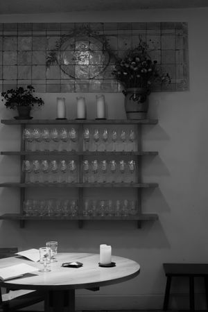 Black and white image of wine glasses displayed on wall shelves with a round table and candle in foreground.
