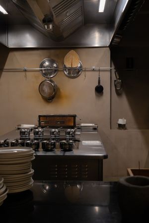 Commercial kitchen setup with stainless steel equipment, plates, and cooking stations under overhead lighting.