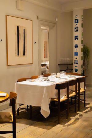 Modern restaurant dining space with white tablecloths, wooden chairs, and minimalist artwork on cream colored walls.