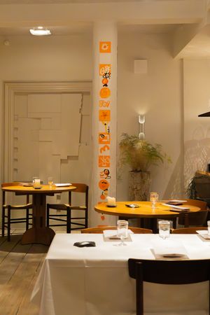 Minimalist dining room with white walls, wooden tables, and orange wall decorations creating an intimate atmosphere.