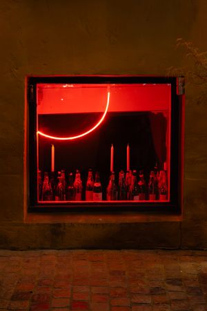 Red neon light installation displaying a curved line above silhouetted bottles in a dark window display.