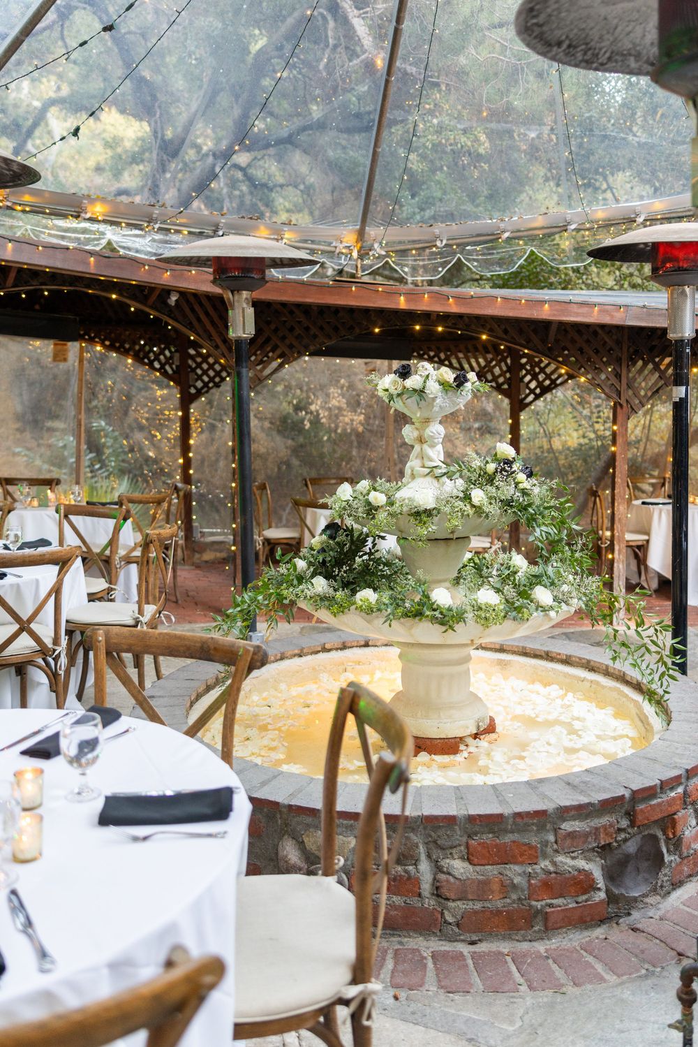 Rustic outdoor wedding venue with fountain centerpiece, twinkle lights, and white floral arrangements under covered patio.