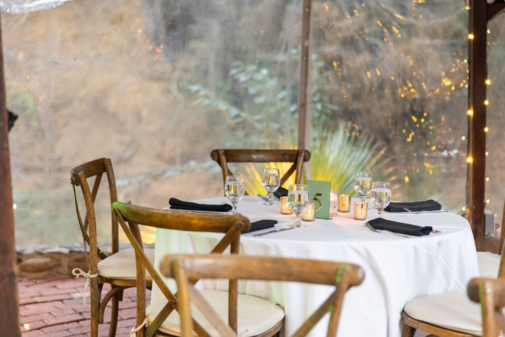 Elegant outdoor wedding reception setup with white tablecloths and wooden cross-back chairs under rustic wooden beams.