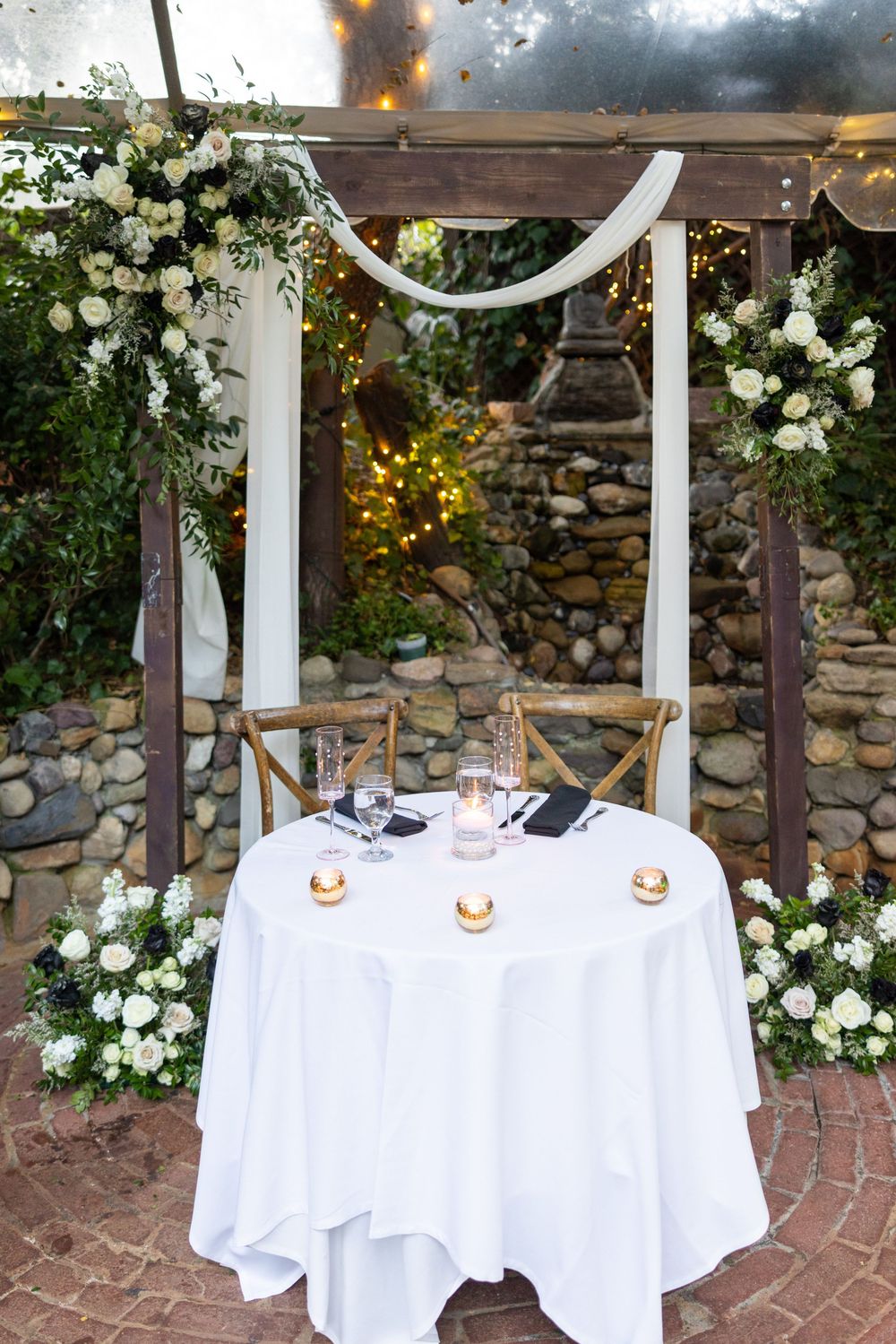 Elegant outdoor wedding ceremony setup with white draped columns, white tablecloths and romantic floral arrangements on stone wall.