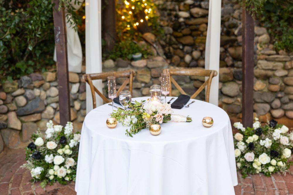 Elegant wedding sweetheart table decorated with white linens, candles and floral arrangements against a rustic stone wall.