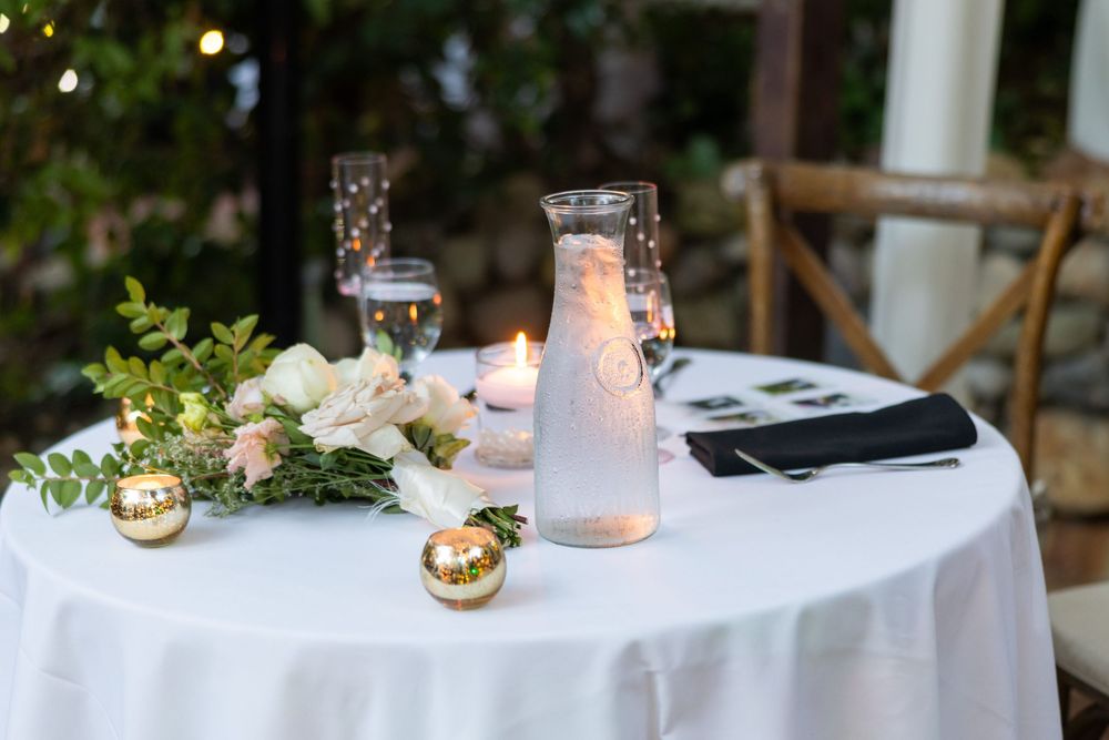 Wedding reception dinner table decorated with white linens, greenery, candles and place settings at dusk.