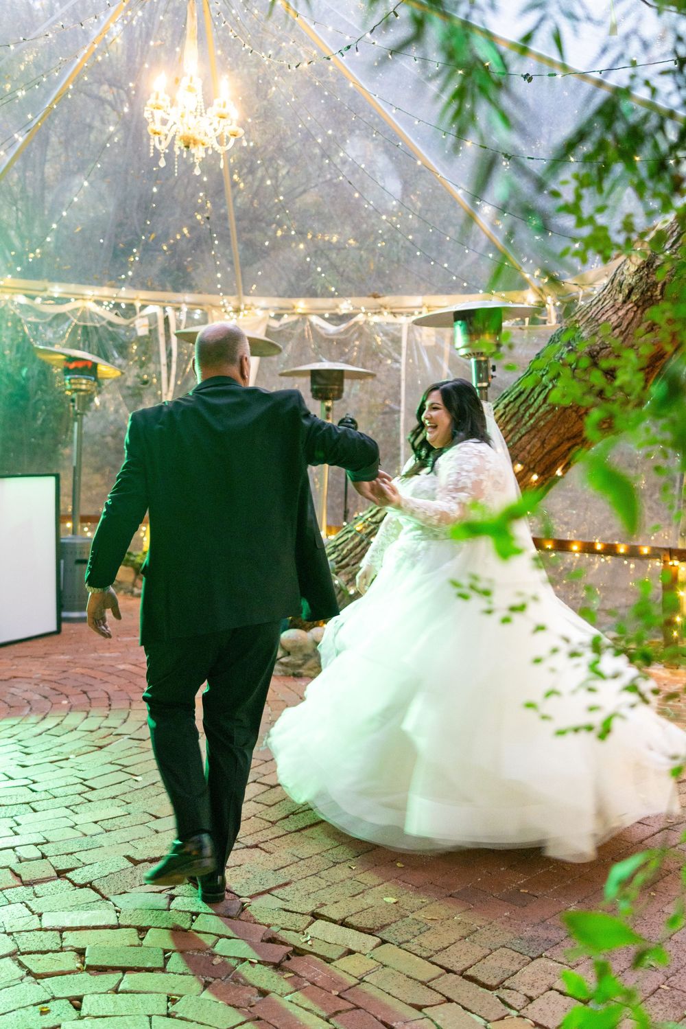 A couple sharing their first dance on a brick patio under an illuminated clear tent with greenery in the foreground.