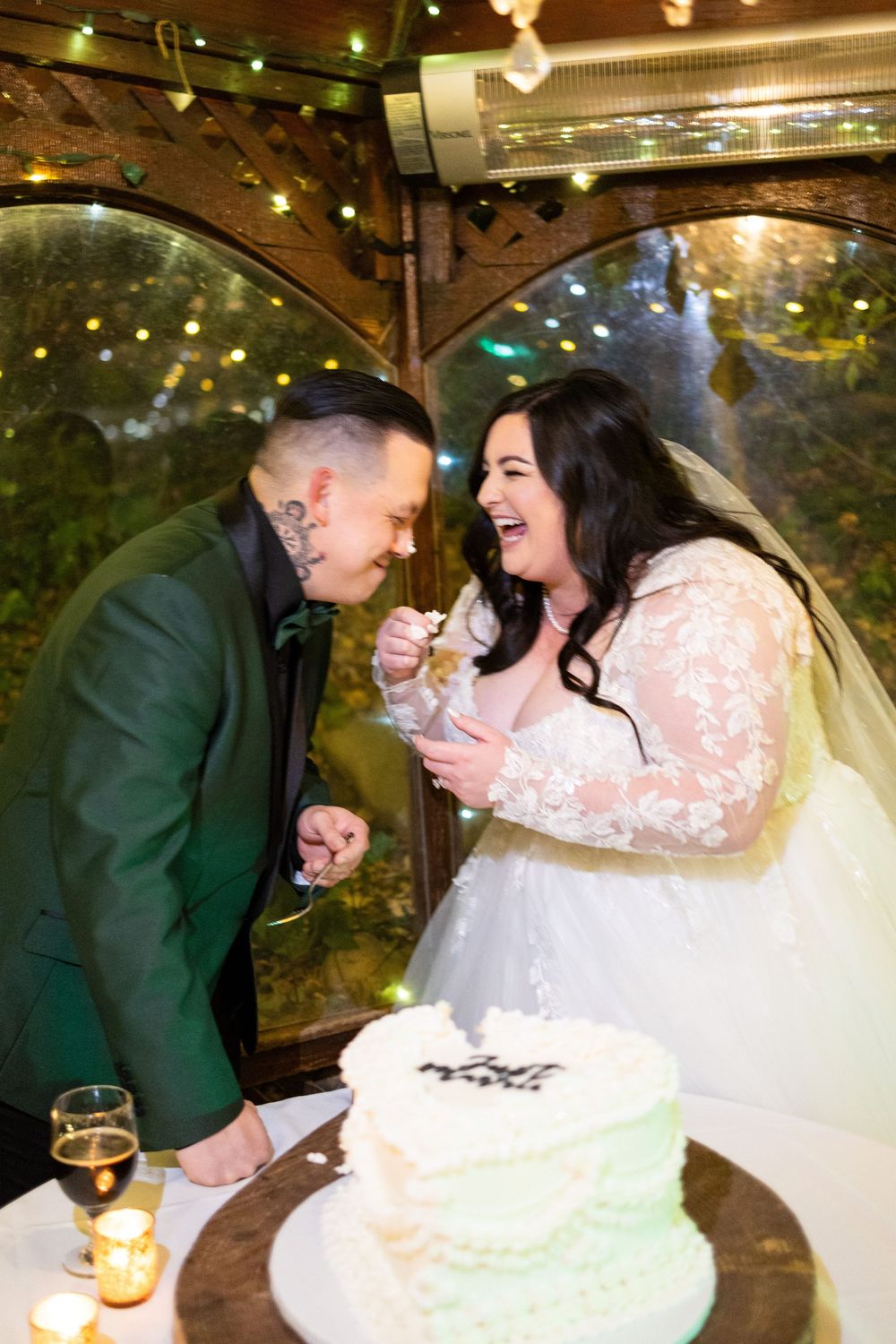 A couple shares a joyful moment while cutting their mint green wedding cake under a wooden arbor with twinkling lights.