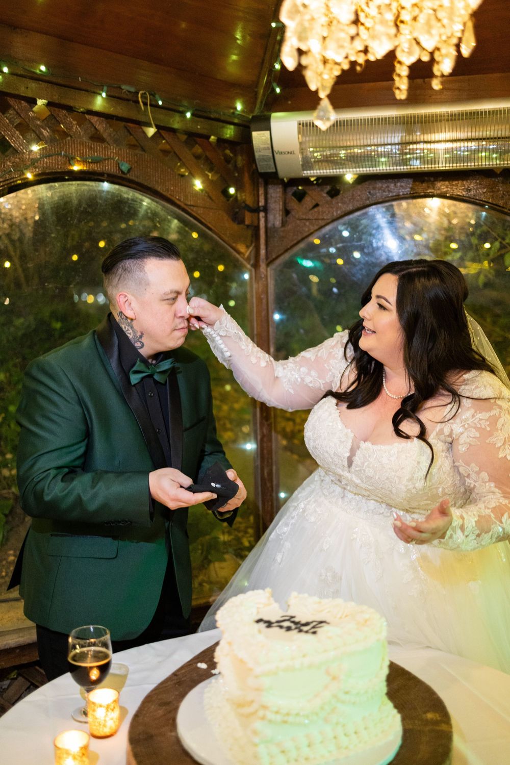 Wedding couple posing with their cake under twinkling lights at an ornate venue.