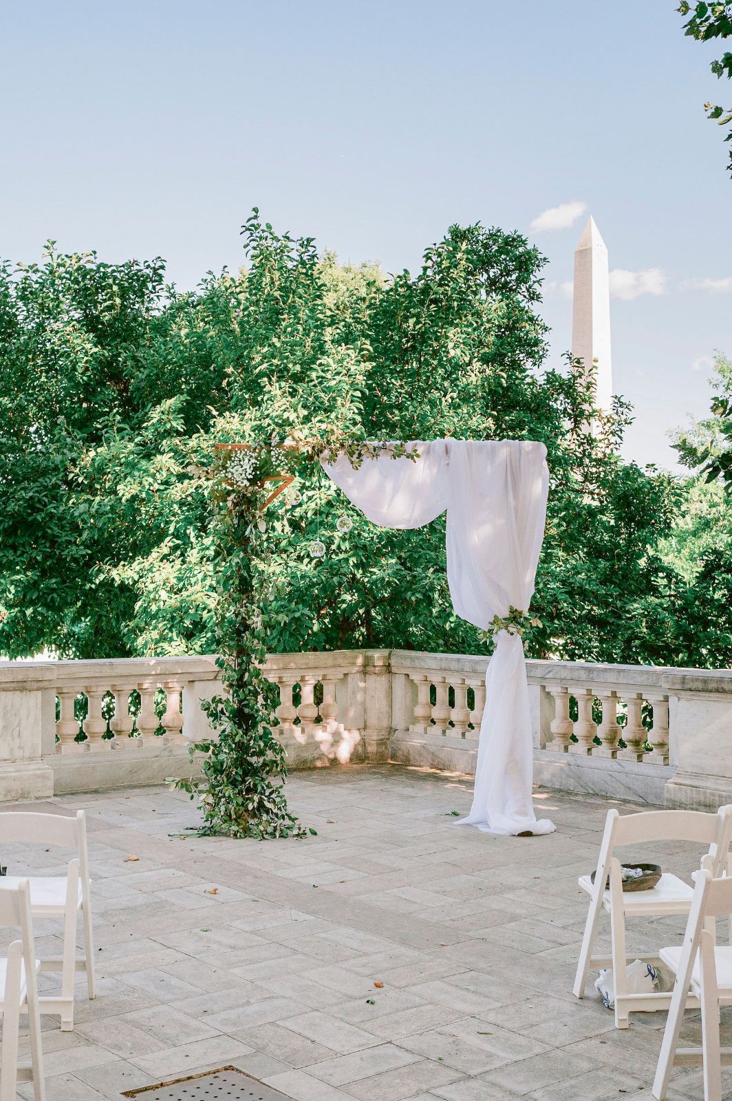 White fabric drapes elegantly over outdoor chairs on a stone terrace surrounded by lush green trees and balustrades.