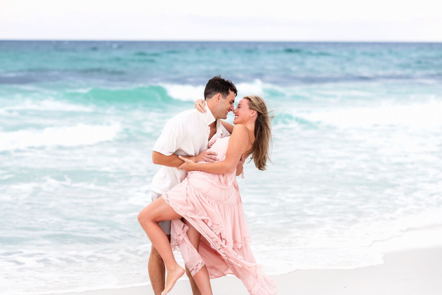Romantic couple dancing playfully together at sunset on a sandy beach with ocean waves in the background.