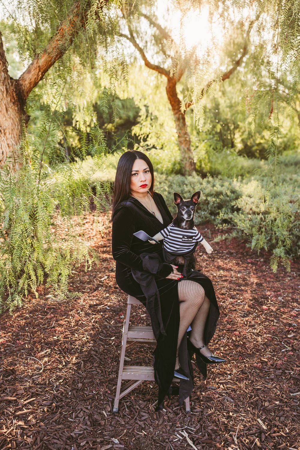 A person in black clothing poses with a small dog under a willow tree in a garden setting.