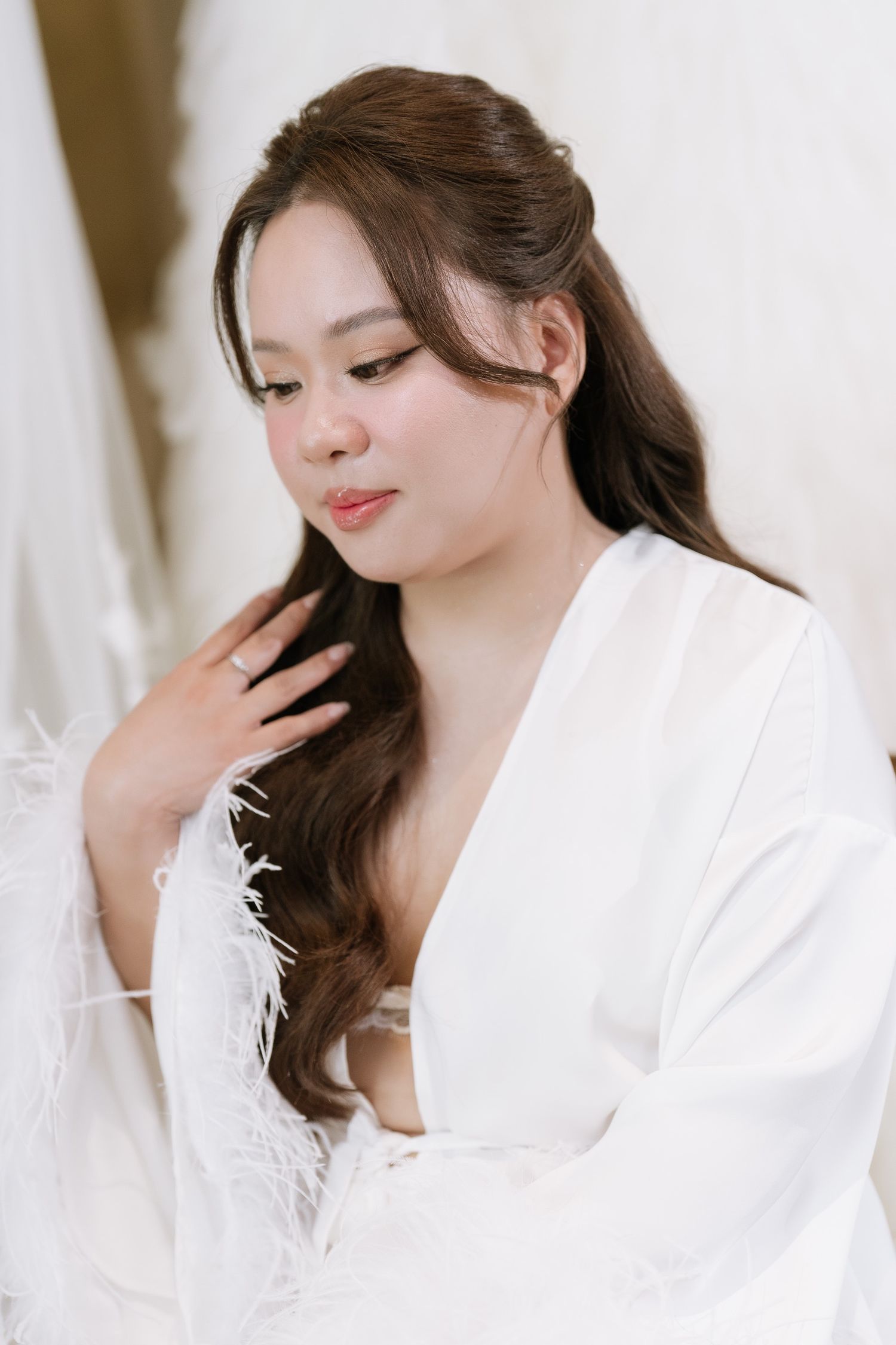 Beautiful bride in white feather robe with soft glam makeup and flowing dark hair looks down gracefully.