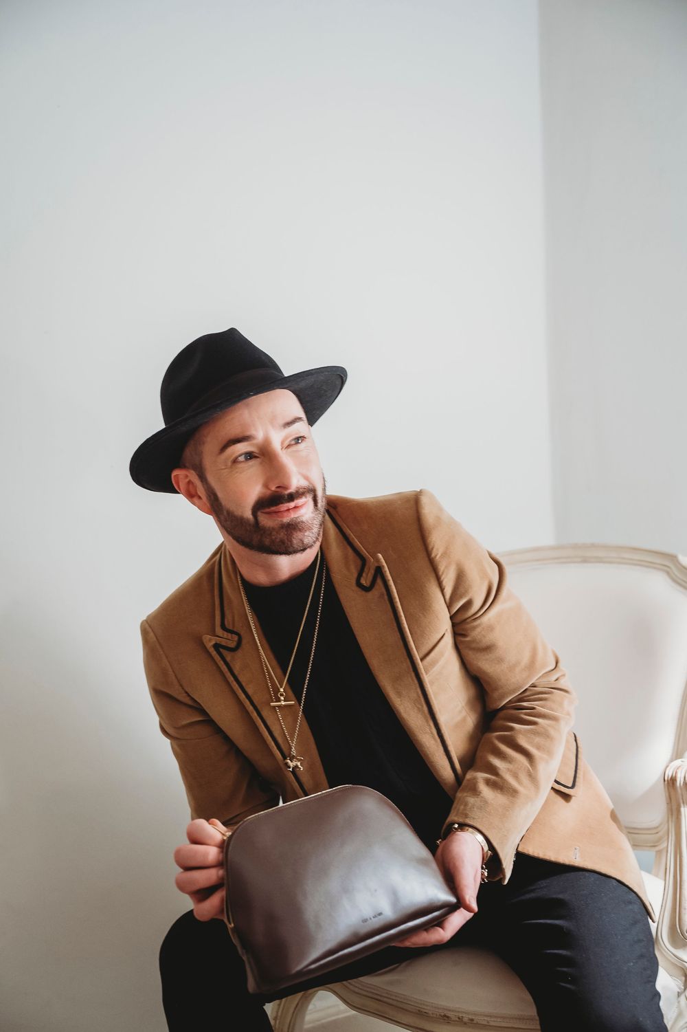 A man in a tan coat and black hat poses against a white wall in a multi-frame photo series.