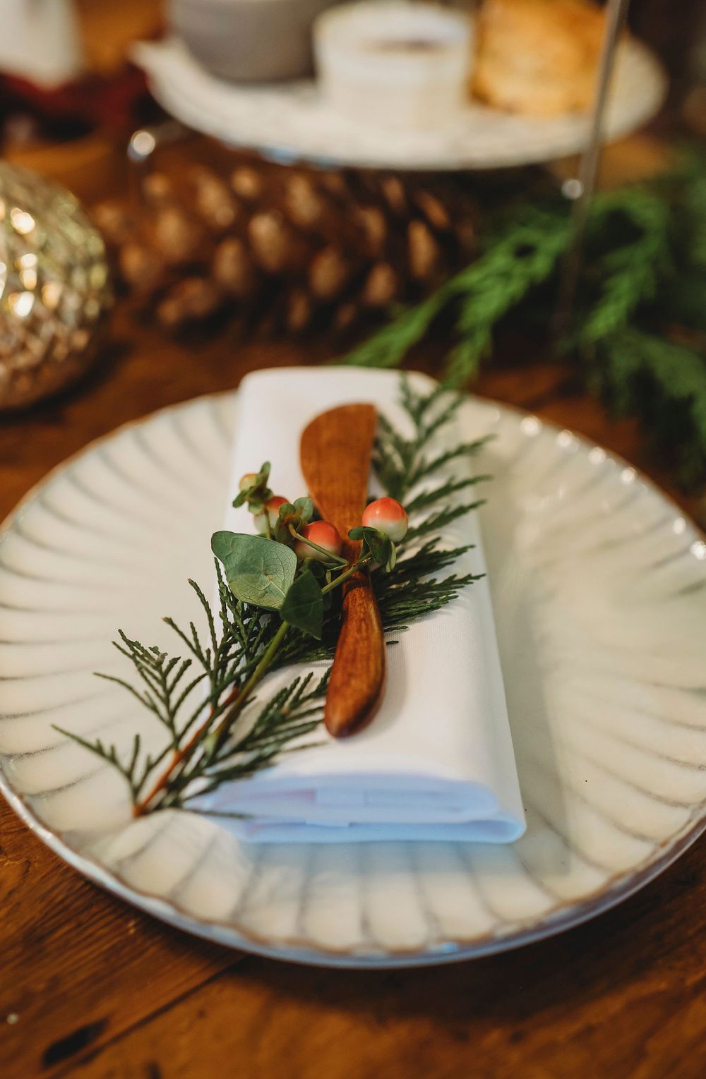 White fluted plates with evergreen sprigs and cinnamon sticks arranged on a rustic wooden table for holiday dining.