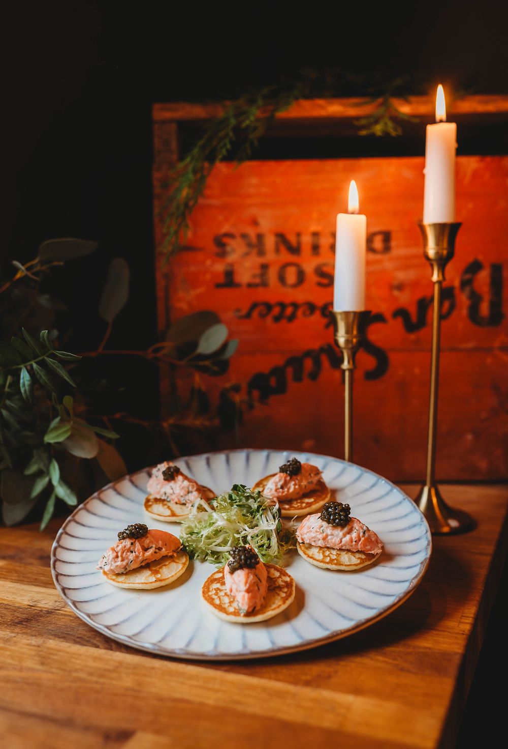 Elegant hors d'oeuvres on a white plate with lit candles in brass holders against a rustic red wooden backdrop.