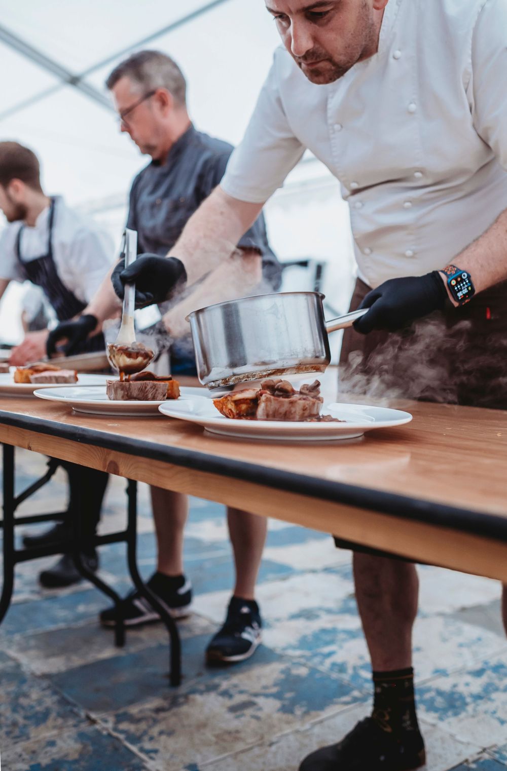 Chefs in white coats plate and garnish dishes on a wooden table over a blue patterned rug.