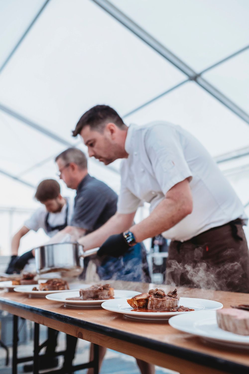 Professional chefs plate gourmet meals at a catered event under a white tent.