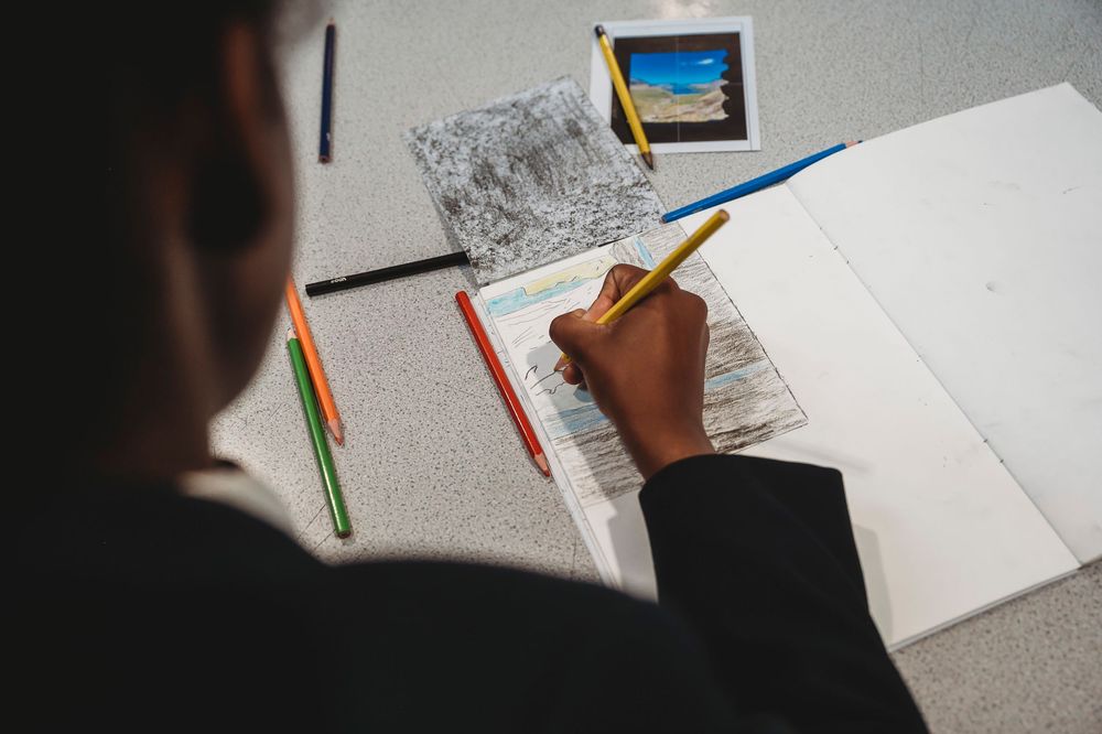 A hand writes in a notebook with colorful pens and papers spread across a white desk surface.