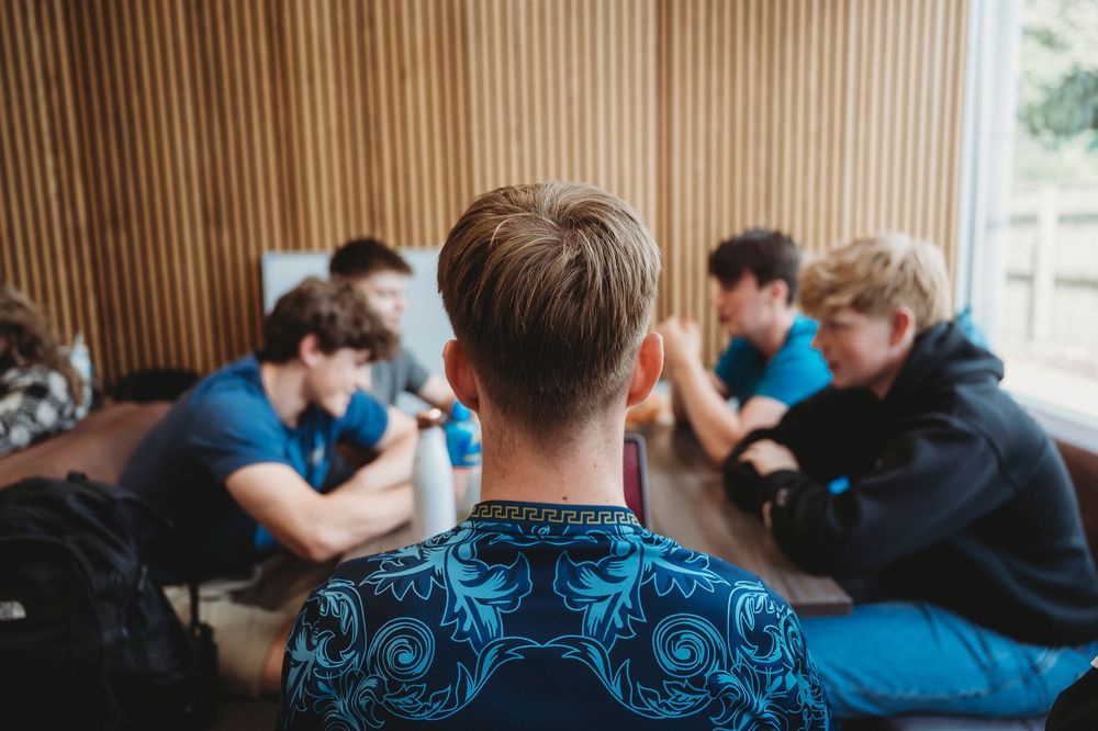 Students sitting at table in study group with one person in blue patterned shirt viewed from behind.