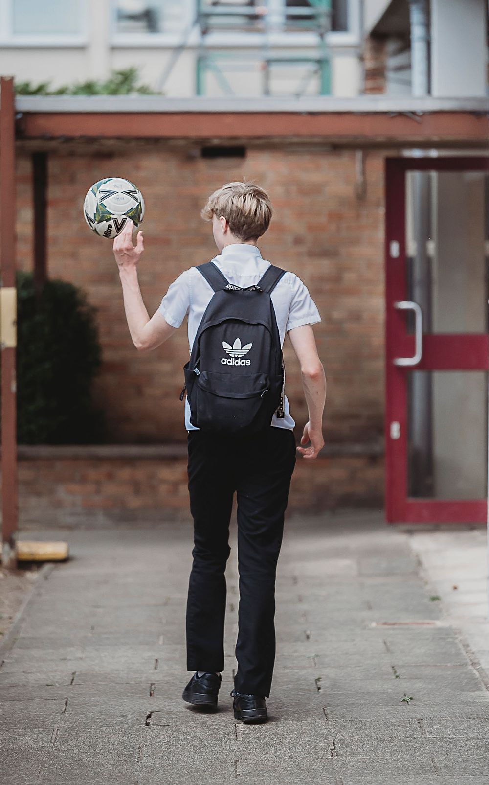 Student wearing an Adidas backpack stands on sidewalk holding a soccer ball in one hand.