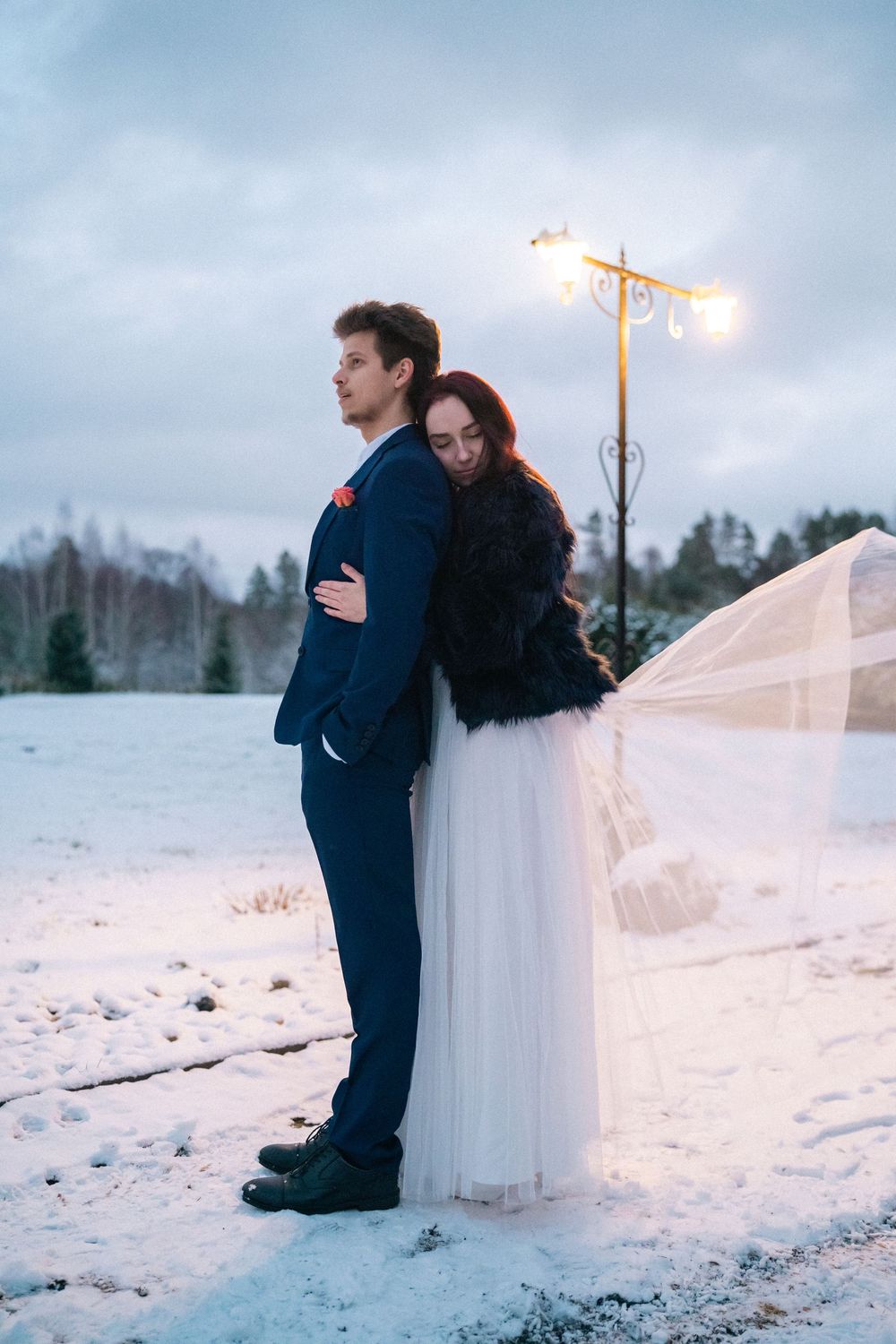 Winter wedding portrait with couple standing in snow at dusk under streetlight.
