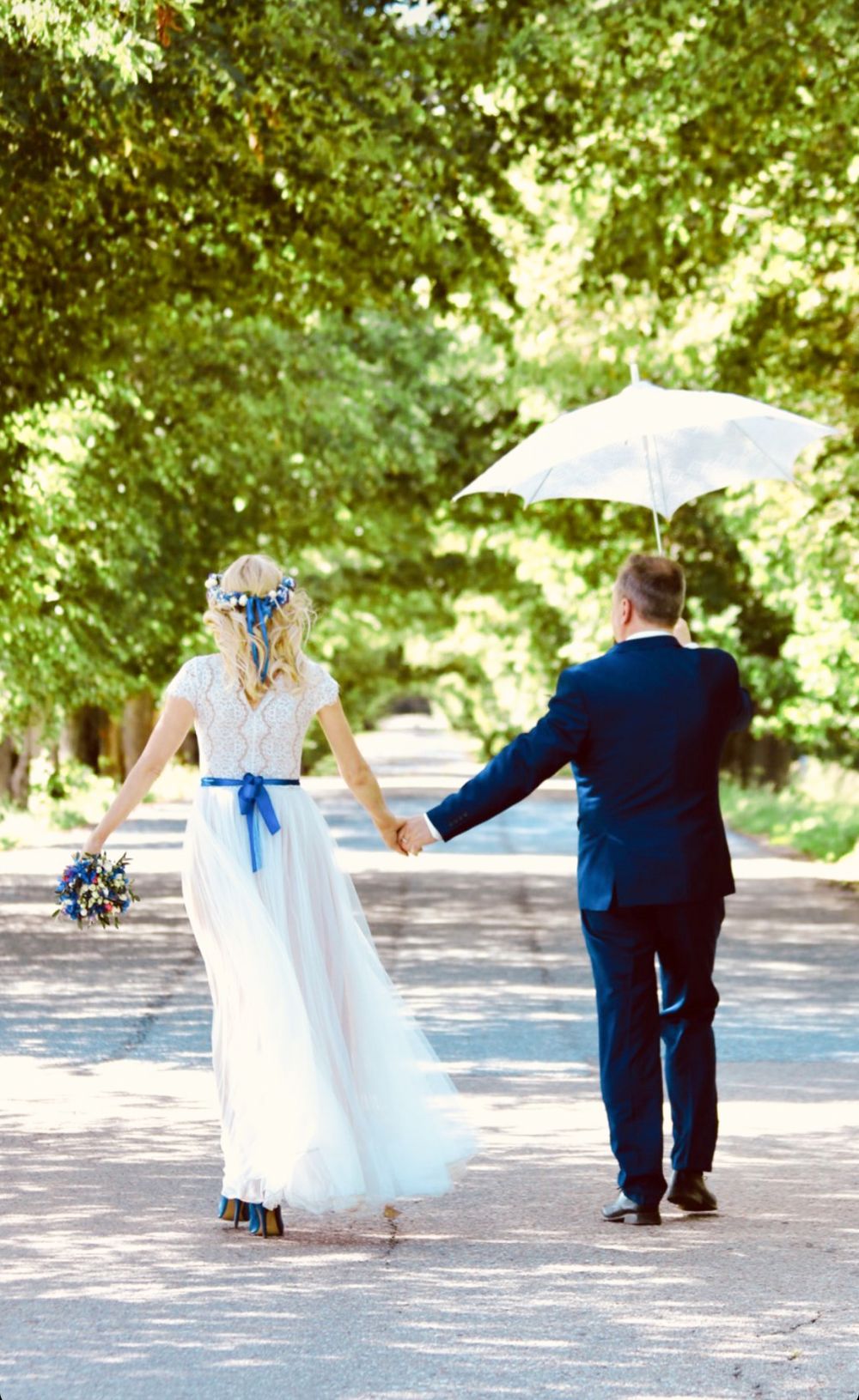 Wedding couple playfully running and dancing together on tree-lined path.