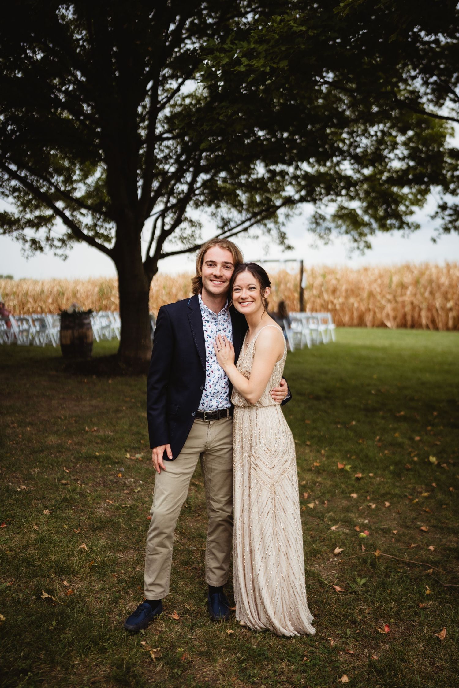 A couple embraces outdoors near a cornfield and large tree on a fall day, with sunlight filtering through the leaves.