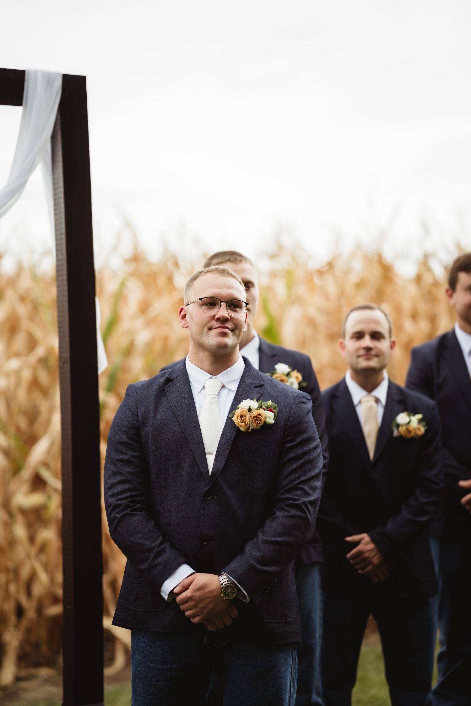 Wedding groomsmen in dark suits stand together in front of a rustic cornfield backdrop at an outdoor ceremony.
