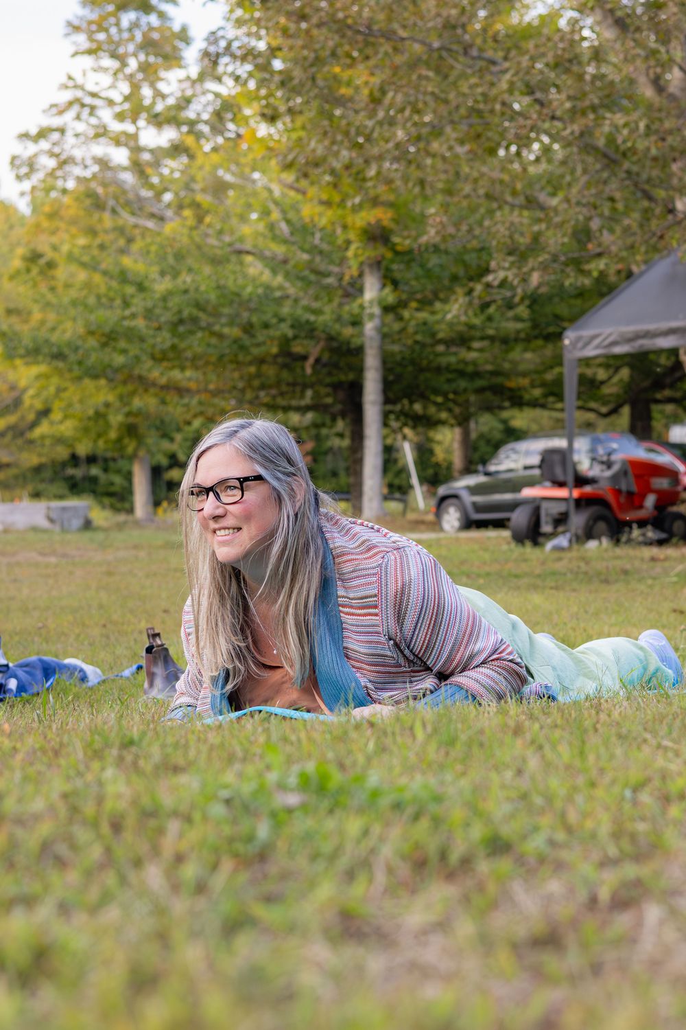 A person lying on grass in a park setting with autumn trees and parked cars in the background.