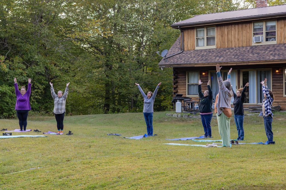 People practicing yoga and stretching exercises together on a grassy lawn near a rustic wooden building.