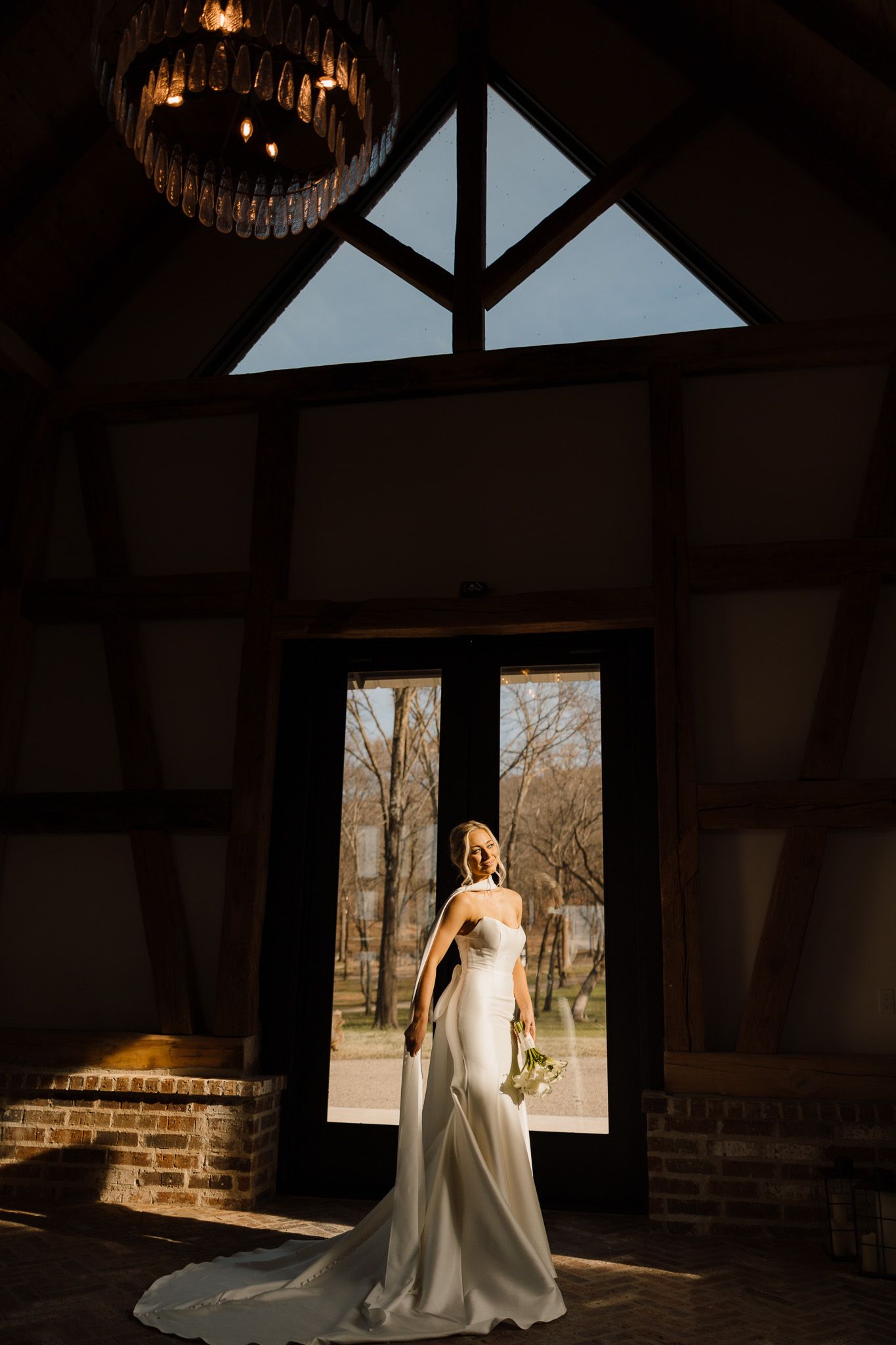 Bride in white gown stands before large French doors in rustic barn venue with dramatic cathedral ceiling and chandelier.