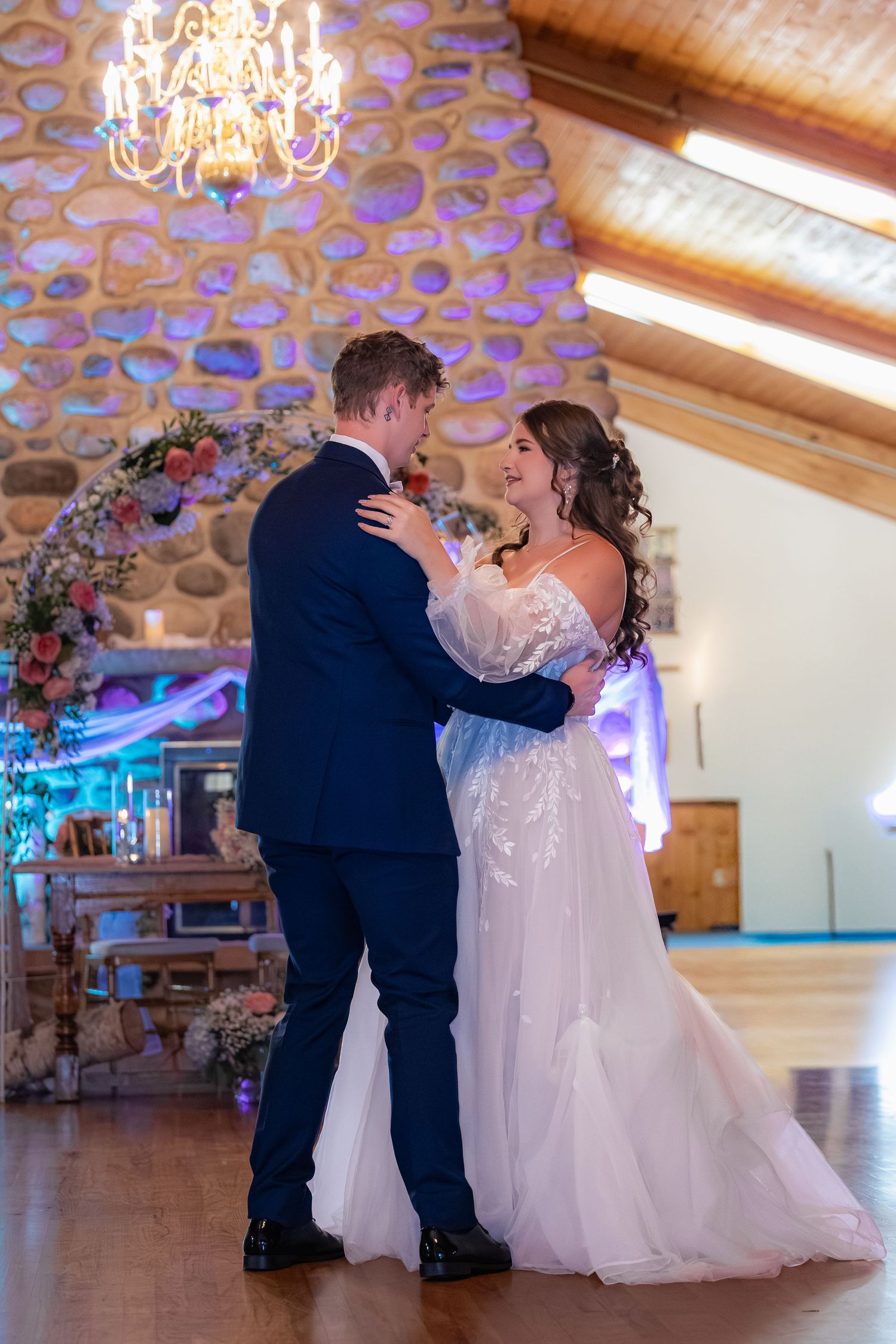 Wedding couple shares their first dance as newlyweds in an elegant ballroom with chandeliers and blue lighting.