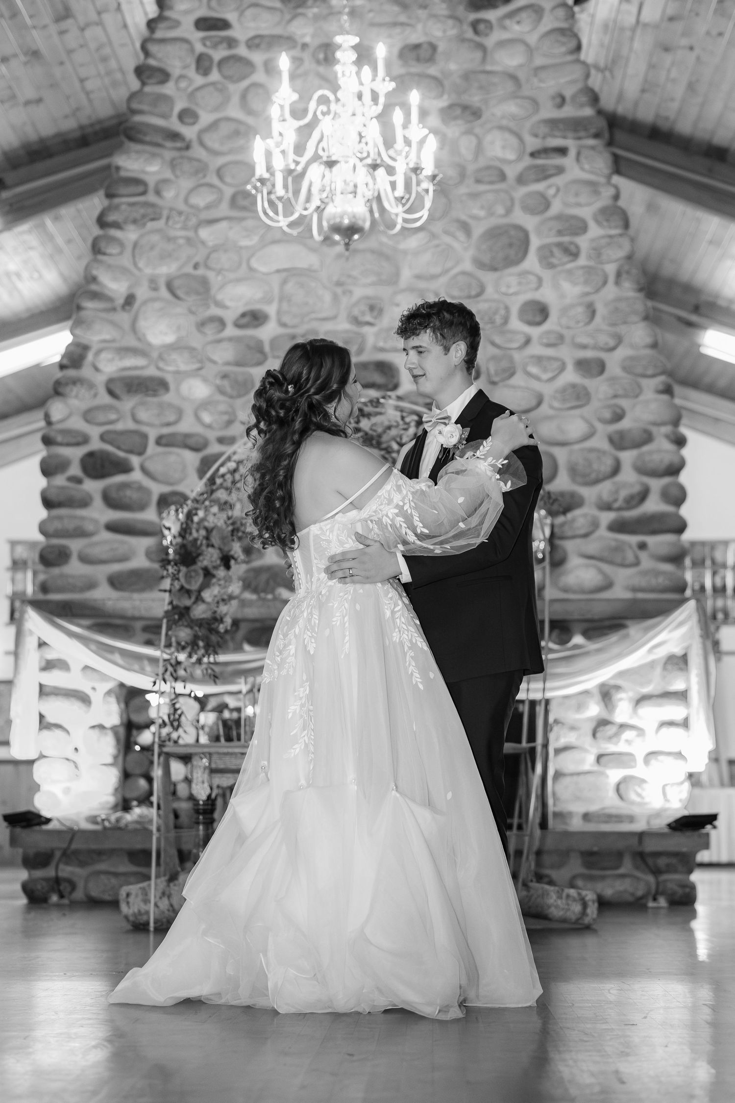 A couple dancing together in elegant wedding attire on a stone wall venue with crystal chandeliers above.