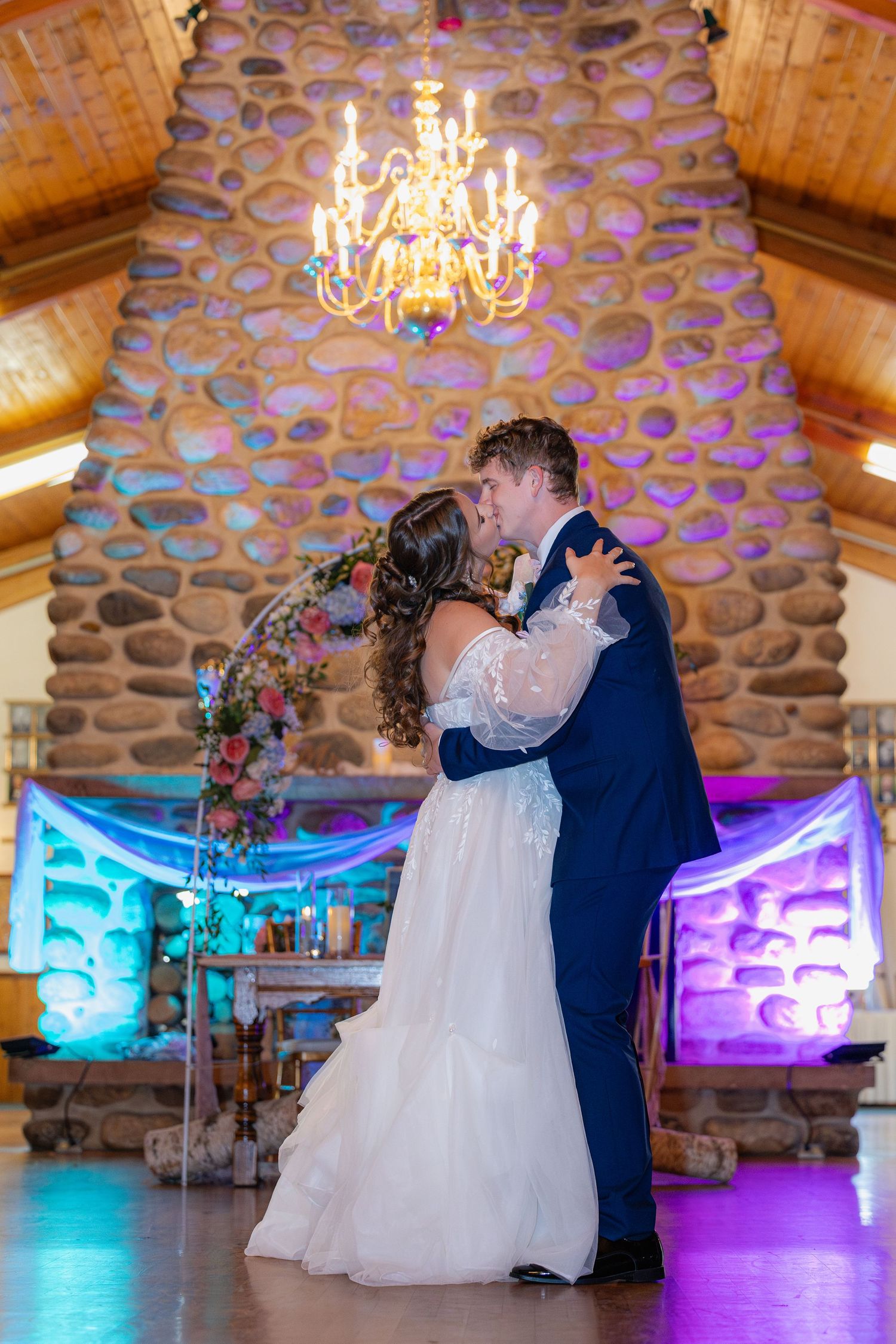 Wedding couple shares romantic dance moments in rustic venue with stone walls and colorful mood lighting.