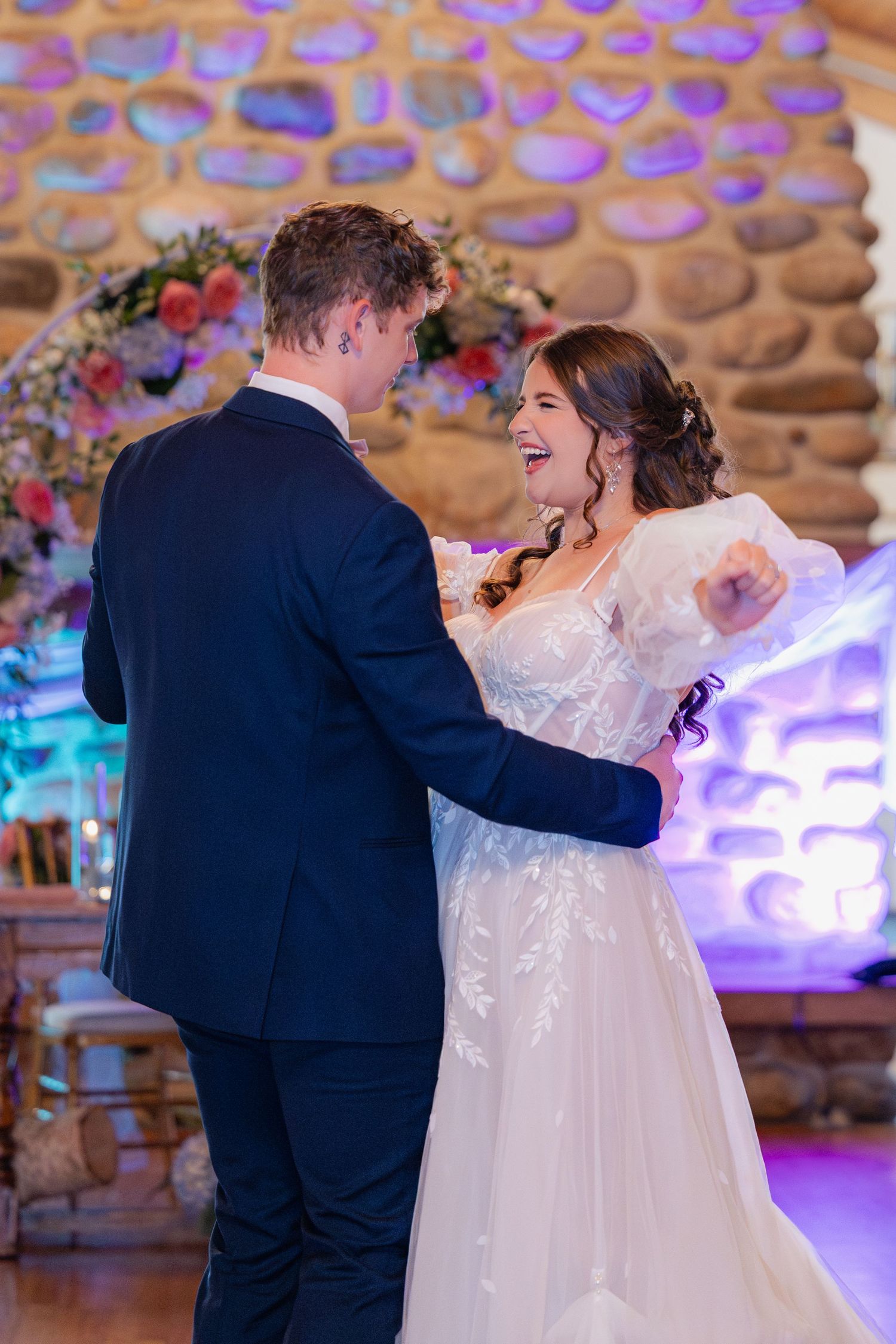 Wedding couple dancing and laughing together under purple ambient lighting in a rustic log cabin venue.
