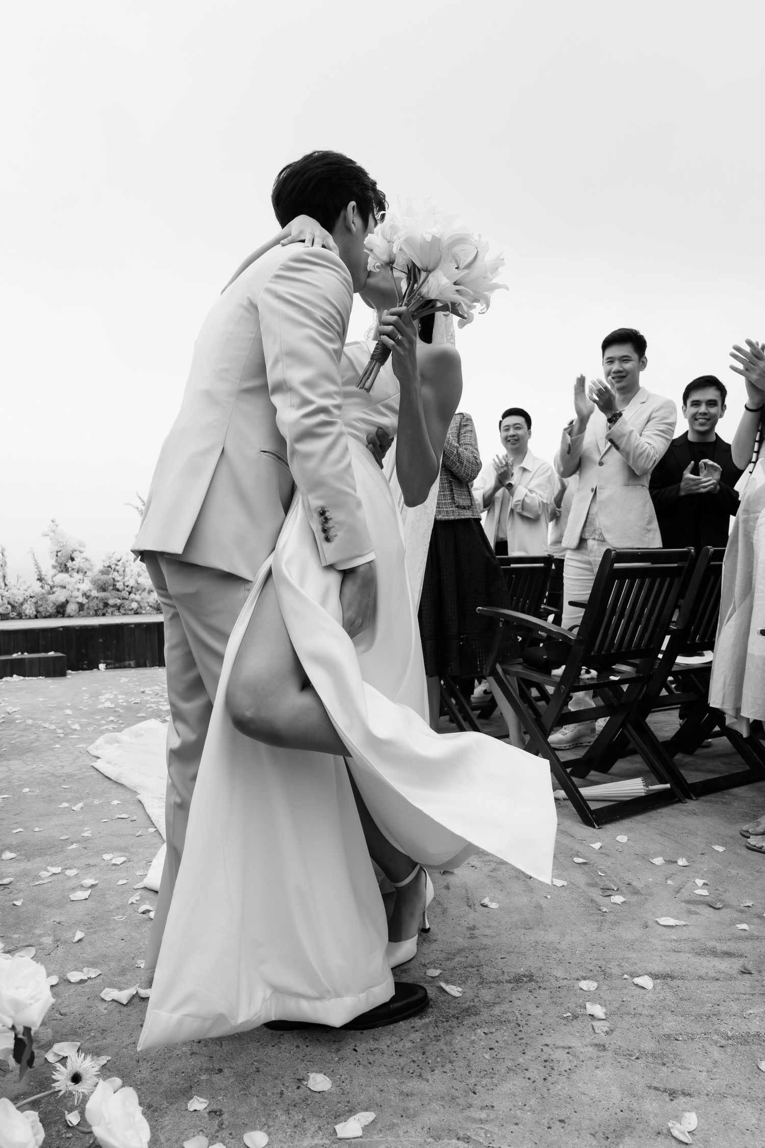Black and white photo of bride and groom sharing first kiss at outdoor wedding ceremony while guests applaud.