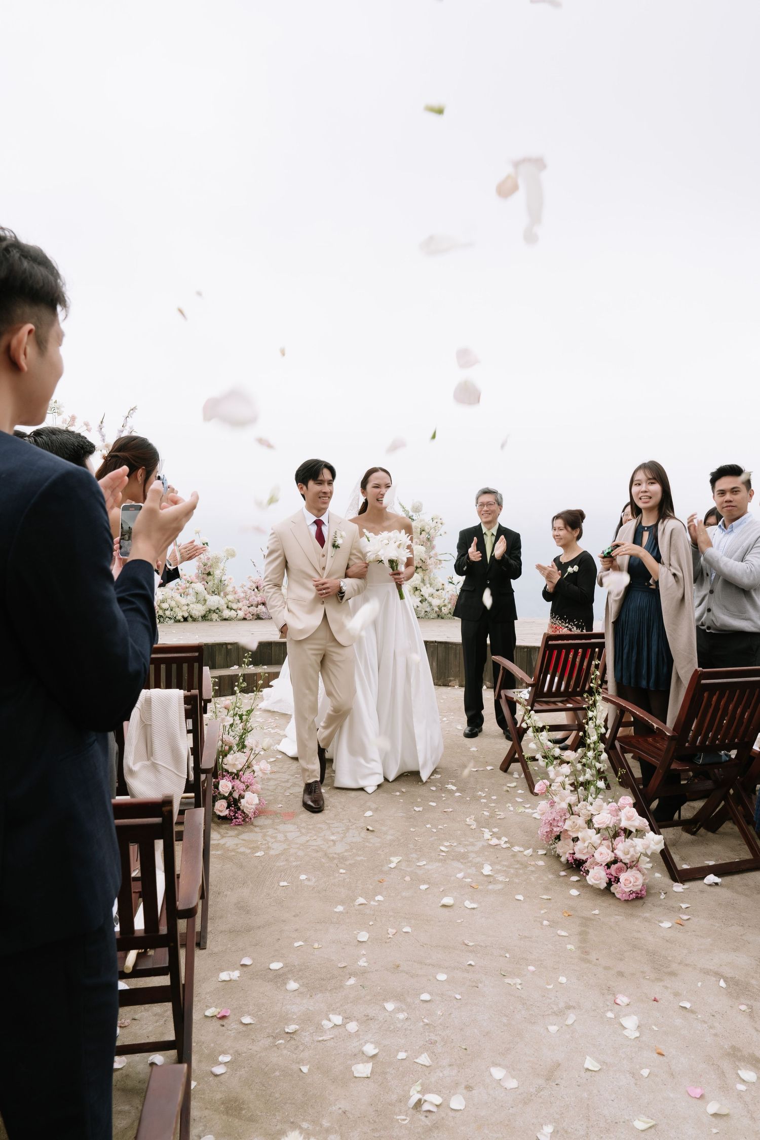 Joyful bride and groom walking down the aisle as guests toss flower petals at an outdoor wedding ceremony.