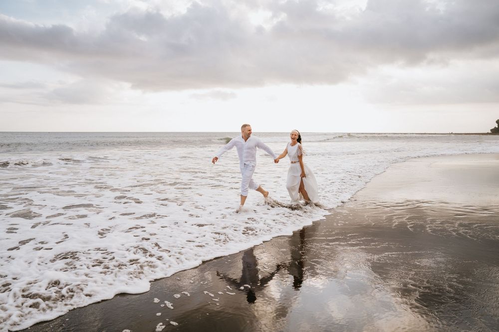 A couple in white outfits walks hand in hand along a dramatic beach shoreline.