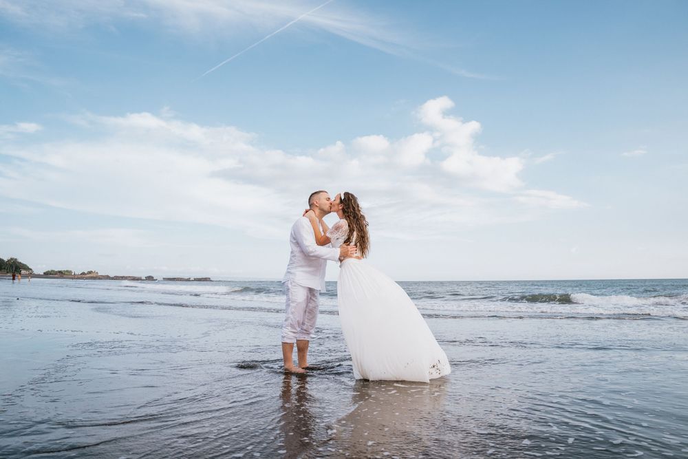 Two people wade in shallow ocean water against a bright blue sky and rocky coastline.