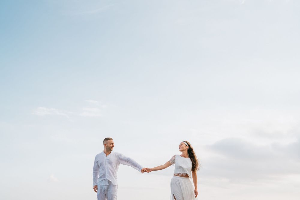 A couple in white clothing walks together on a serene beach during sunset.