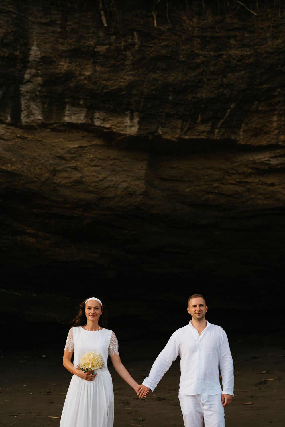 A couple in white attire holds hands while standing beneath a dramatic rocky overhang at sunset.