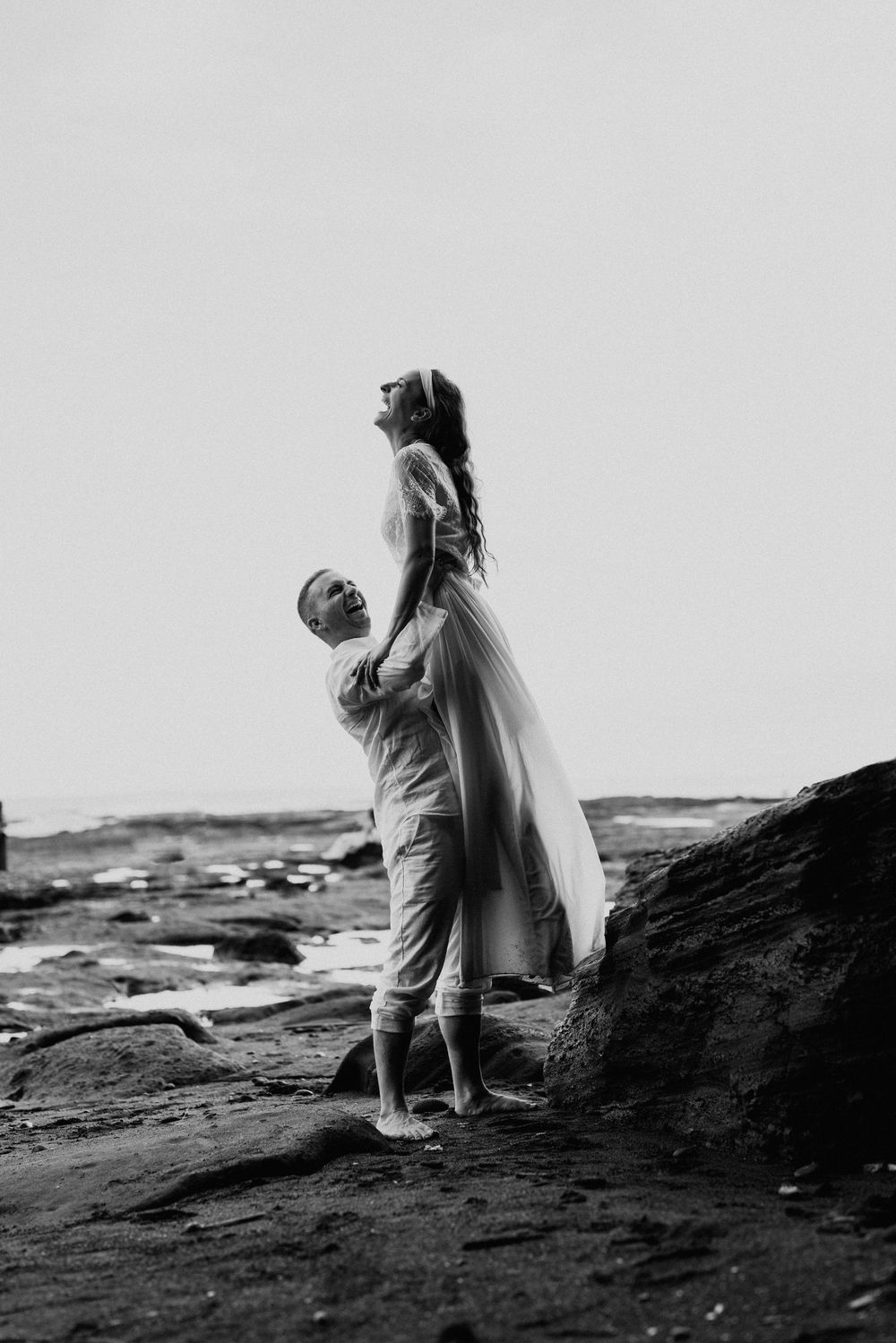 A romantic black and white photo of a couple embracing on rocky coastal terrain.