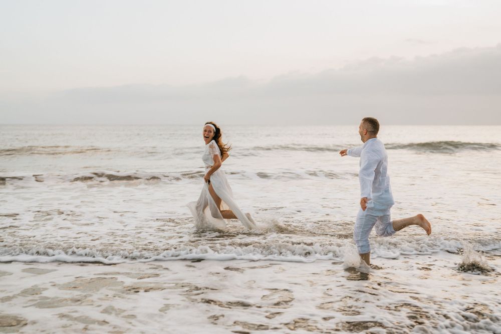 A couple runs playfully along the beach at sunset with waves crashing nearby.