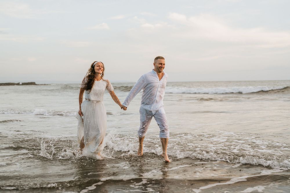 A joyful couple splashes through shallow ocean waves in matching white outfits.