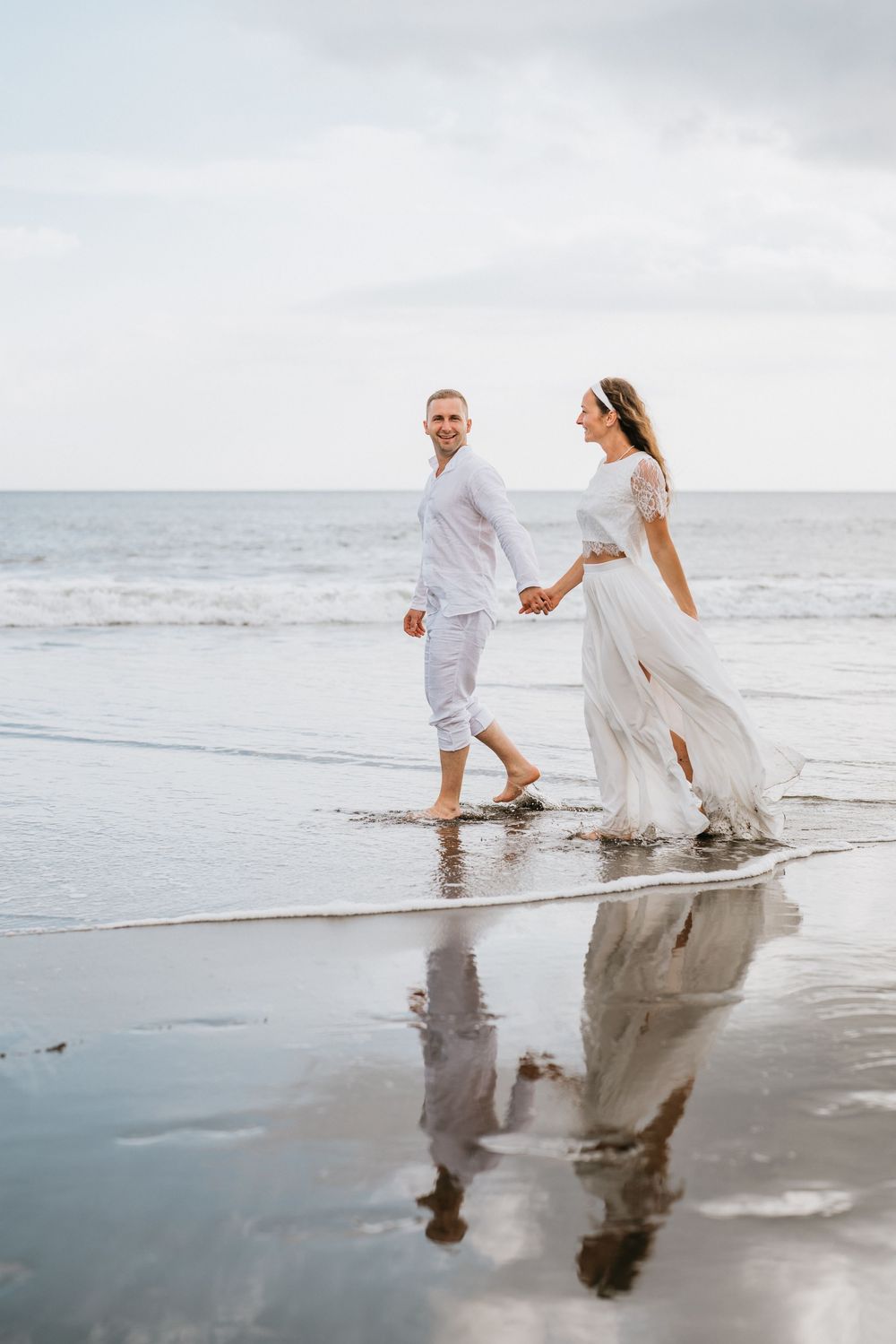 Two people in white clothing walk along a reflective beach shoreline at dusk.