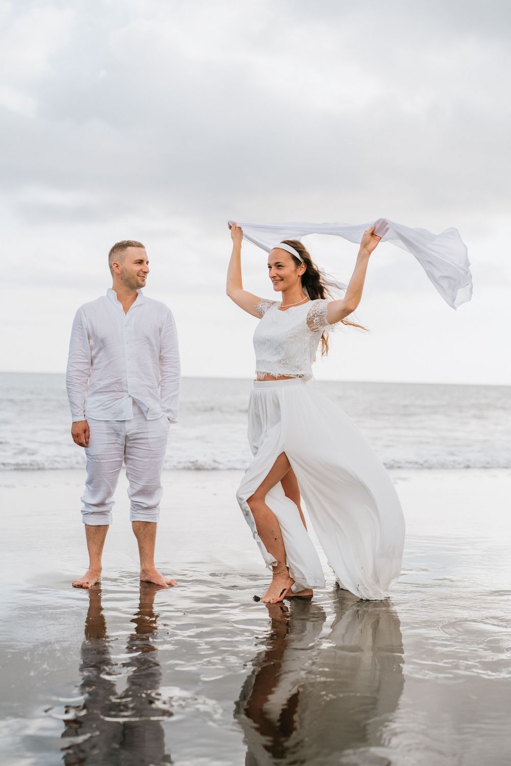 A couple in white outfits plays in shallow ocean water during an overcast beach photoshoot.